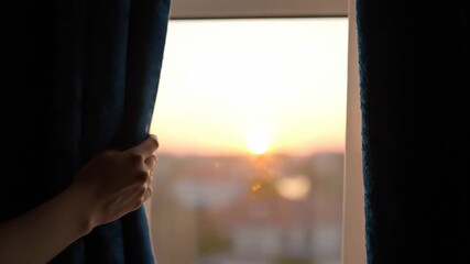 Delicate hands parting deep blue curtains at the break of day, as the sun's rays illuminate a stunning sunrise over a tranquil urban landscape