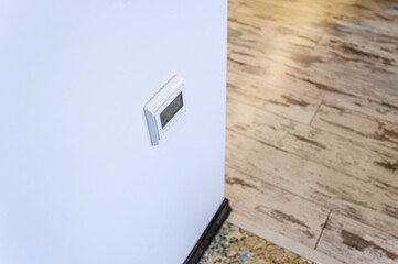 A white thermostat is mounted on a white wall, with wood-patterned flooring visible in the background