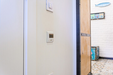 A bright interior shot focusing on a wall with a light switch and a thermostat. Partially visible is a wooden panel and a glimpse into an adjacent room
