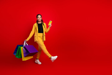 Young stylish woman in vibrant yellow outfit holding colorful shopping bags while smiling confidently against a bold red background.