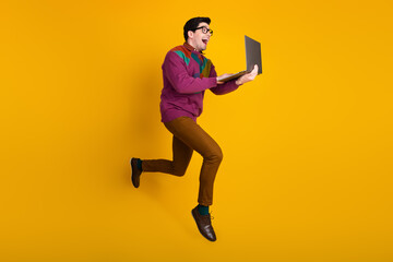 Happy young man in colorful sweater jumping energetically holding a laptop over a vibrant yellow background