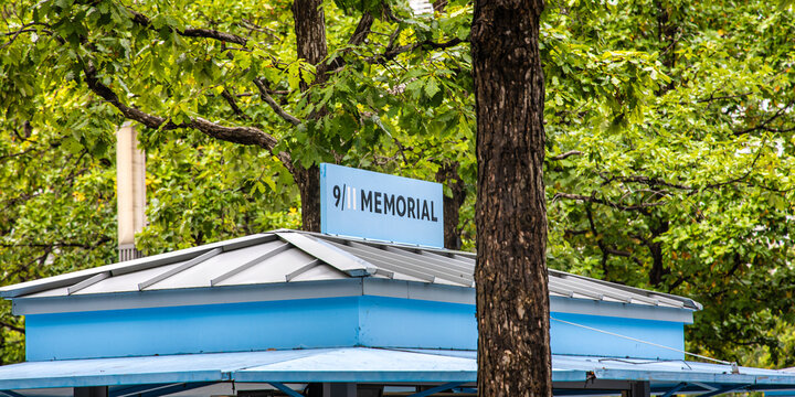 Blue 9/11 Memorial sign atop kiosk surrounded by green trees in New York City