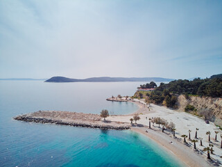 Aerial view of a serene coastal landscape with a sandy beach, clear blue water, and a rocky pier extending into the sea. The shoreline is lined with palm trees, and distant hills are visible.