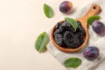 Dried prunes in a wooden bowl with fresh plums and green leaves on a light beige background
