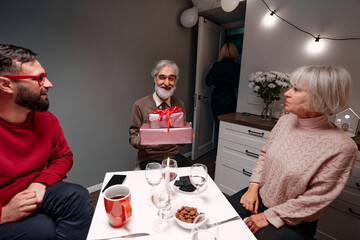 Elderly man receiving a wrapped gift, surrounded by family, fostering joy and togetherness. Depiction of family celebrations, warmth, and appreciation in a welcoming and festive indoor setting.