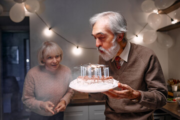 An elderly man blowing out candles on a birthday cake while a woman watches excitedly in a festive home setting with balloons and warm lighting, conveying joy and togetherness.