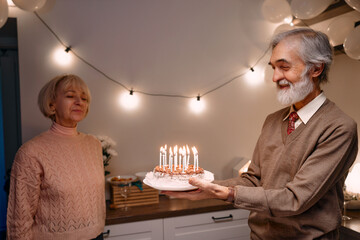 Joyful moment of senior couple celebrating birthday with candle-lit cake. Decorated surroundings with fairy lights and balloons enhance the festive ambiance, highlighting their bond and happiness.