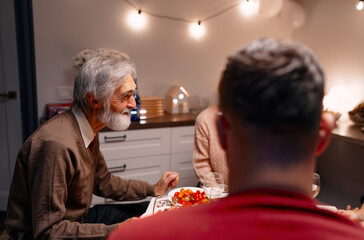 An elder and family members are gathered around a well-set table enjoying a meal in a cozy home adorned with warm lighting and a festive environment, evoking warmth, togetherness, and celebration.