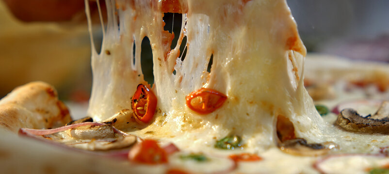 Delicious pizza slice being pulled from a large, fresh, and hot pizza, creating long, gooey strands of molten mozzarella cheese, garnished with tomatoes and mushrooms