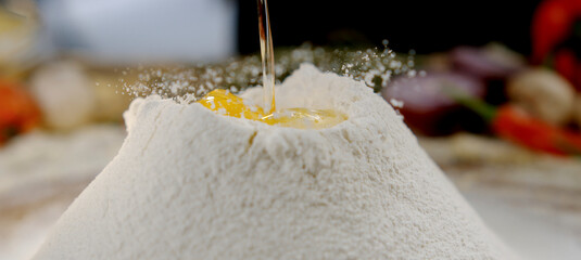 White wheat flour mound on a wooden table formed into a well, receiving a raw egg while golden olive oil pours in for homemade pasta or rustic bread dough preparation