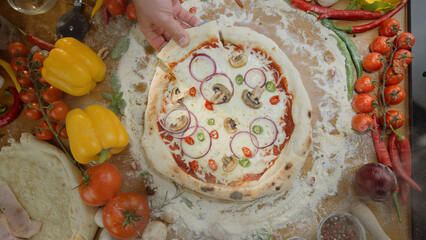 Person's hand taking a slice of homemade mushroom, onion, and pepper pizza from a rustic wooden board, surrounded by fresh vegetables like tomatoes and bell peppers