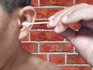 A man holding a cotton swab is cleaning his left ear on a brown brick wall background.