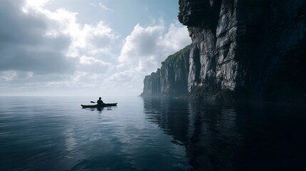 A lone kayaker paddles on a calm sea beneath dramatic cliffs under a cloudy sky