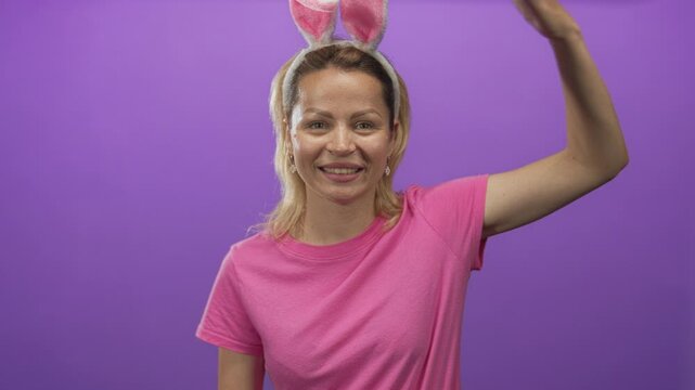 Young blonde woman raises hand in studio wearing bunny ears headband and pink t shirt while smiling with closed eyes; playful joy.