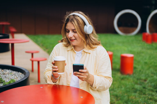 Happy young Caucasian woman in good mood listening to music with headphones and sitting on terrace street cafe in the city. Music lover enjoying music. Urban lifestyle concept.