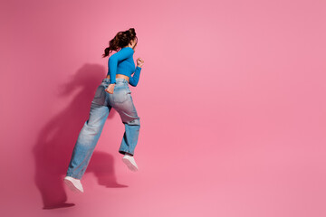 Energetic woman in casual outfit jumping freely against pink background, full-length profile view showcasing dynamic posture