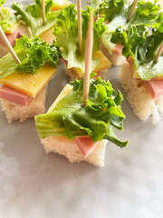 Small canap&eacute;s made from square pieces of bread, salami, hard cheese, and fresh lettuce, held together with a wooden toothpick, are arranged on a gray textured plate. Close-up. Appetizer. Buffet.