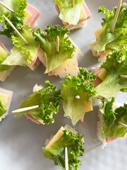 Small canap&eacute;s made from square pieces of bread, salami, hard cheese, and fresh lettuce held together with a wooden toothpick are arranged on a gray textured plate, close-up, top view. Appetizer.
