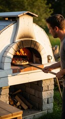 Obraz premium Man Baking Pizza in Outdoor Wood-Fired Oven at Sunset.