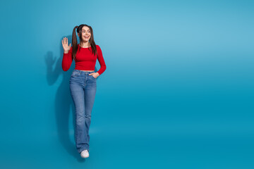 Cheerful young woman waving hello against a vibrant blue backdrop wearing casual red top and jeans © deagreez
