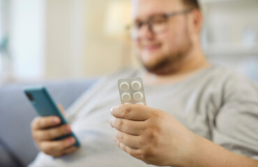 Obesity diet overweight man with pills smartphone. He compares medication details online at home, weighing health risks and nutrition advice. Clear concept: mindful weight control choices.