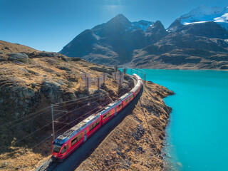 Aerial view of red Bernina Express train passing by turquoise lake Bianco in Swiss Alps, Switzerland in autumn. Scenic railway route with mountains, lake, and train journey in fall. Top drone view