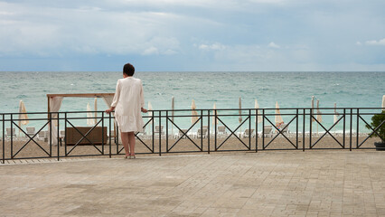 A stroll along the seafront promenade. A solitary figure of an adult woman in a white dress stands by the railing against the backdrop of the sea, a beach umbrella, and sun loungers.