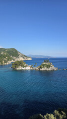 Fototapeta premium Small island with church and pine trees in Ionian Sea near Parga Greece. Scenic seascape with blue water, mountains and sky. Popular travel landmark and Greek coastal view.