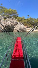 Fototapeta premium Red-carpeted boat ramp leading down into the crystal clear, shallow turquoise water toward a rocky, tree-covered cove in Adrasan