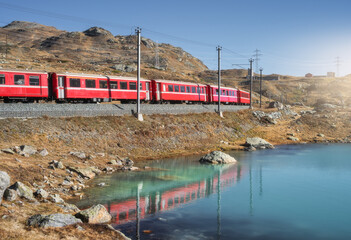 Bernina Express train near lake in Switzerland at sunset, clear reflections, mountain silhouettes and alpine terrain in peaceful evening light in the Swiss Alps. Red train. Transport. Railway station