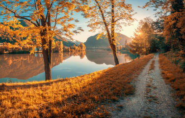 Scenic autumn view of a river with mountains reflected in calm water. Trees with golden foliage line a countryside path at sunset in Slovenia. Vibrant seasonal colors and peaceful nature in fall