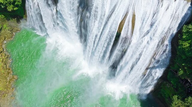 Aerial shot of a spectacular and powerful waterfall cascading into a beautiful turquoise plunge pool surrounded by lush green forest.