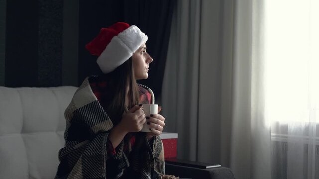 Cinematic shot of a young sad woman in a Santa hat sitting at home wrapped in a blanket and holding a cup of hot drink. Lonely Christmas mood, emotional and reflective moment, winter melancholy