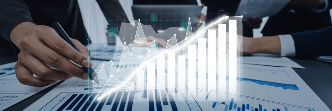 A business team collaborates in a modern office space to analyze financial data and trends. The image features detailed charts and a digital overlay, showcasing growth. Axiom - Powered by Adobe