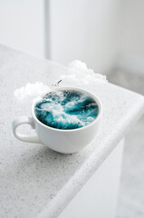 Surreal photo manipulation of a white coffee cup filled with ocean waves, clouds, and a flying seagull. Conceptual fantasy artwork.