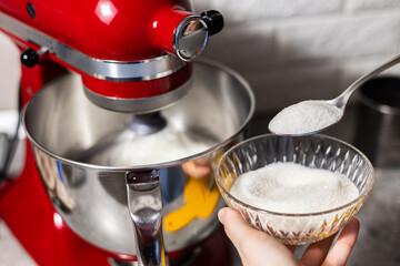 Sugar in spoon is poured into the bowl of a planetary mixer with beaten eggs.