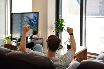Young man sitting on a couch at home watching American football on television with raised arms celebrating a play