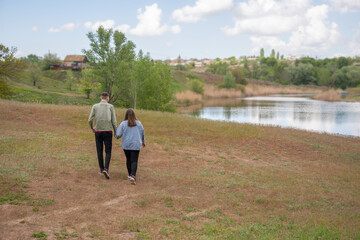 Young couple, a boy and a girl, stroll hand in hand along the grassy bank of a pond, symbolizing love, romance, partnership, and spending time together in nature