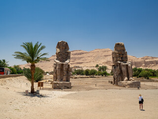 LUXOR, EGYPT : The Colossi of Memnon, two massive stone statues of Pharaoh Amenhotep III, stand on the west bank of the Nile, surrounded by desert landscape under a clear sky.
