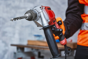 Close-up of a worker in safety vest and gloves holding a red electric drill with a drill bit for construction or renovation work. Concept of power tools, safety and professional equipment.