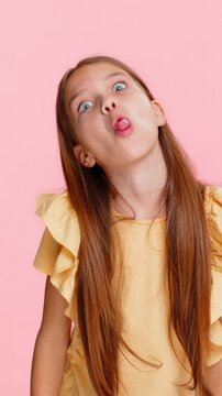 Young child girl sticks out tongue teasingly while making goofy faces cheerful grimaces into camera. School kid isolated on pink background, acting silly and playful with joyful gestures and emotions