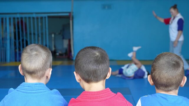 Three young boys in sambo uniforms watch a sports match on the mat, with a referee in the background giving instructions. Concept of martial arts, discipline, and children sports.