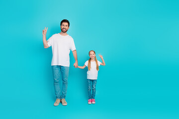 Fototapeta premium Dad and daughter wearing white t-shirts showing peace gesture against a blue studio backdrop, displaying joy