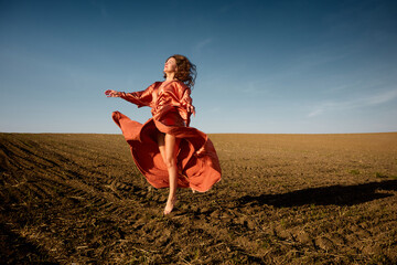 A captivating image features a woman in a flowing red dress dancing barefoot in a sunlit field...