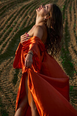 Fototapeta premium A beautiful young woman dressed in an orange dress, she stands in a vast plowed field, with her face turned upwards, enjoying the warmth of the sun and the fresh air, feeling the serenity of the