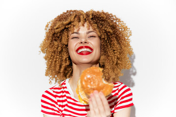Happy woman with curly hair enjoying a burger, wearing a striped shirt and bright red lipstick, representing joyful eating and indulgence in fast food