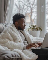 A young Black man with curly hair sits on a couch, wrapped in a white blanket, working on a laptop in a cozy home office during winter.