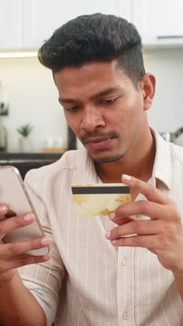 Indian man at home orders delivery on smartphone enters card data checks address and confirms payment. Arabian guy on sofa shows thumbs up approval order confirmed waits courier tracking notification