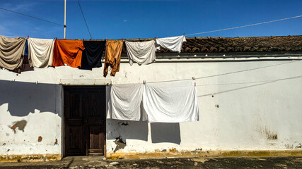 Laundry hangs on two clotheslines against a sunlit white wall with an old wooden door and terracotta roof. Features architectural heritage and sustainable domestic living