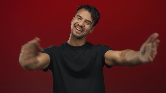 Young hispanic man in a black shirt smiles warmly while embracing himself against an isolated vibrant red background.